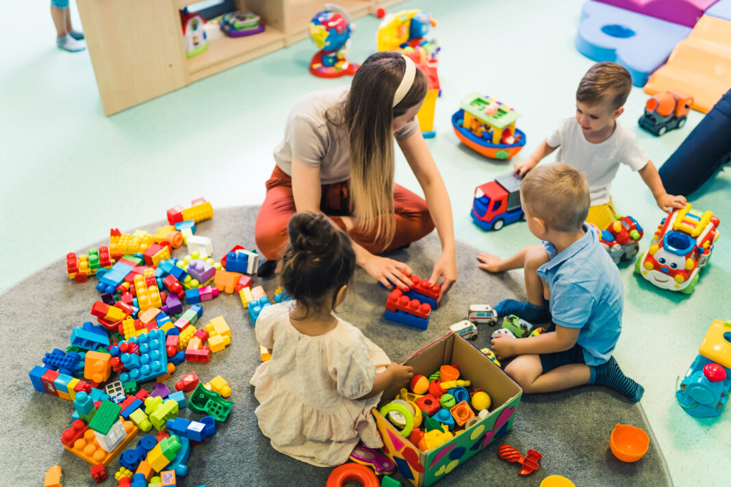 Playtime at nursery school. Toddlers with their teacher sitting on the floor and playing with building blocks, colorful cars and other toys.