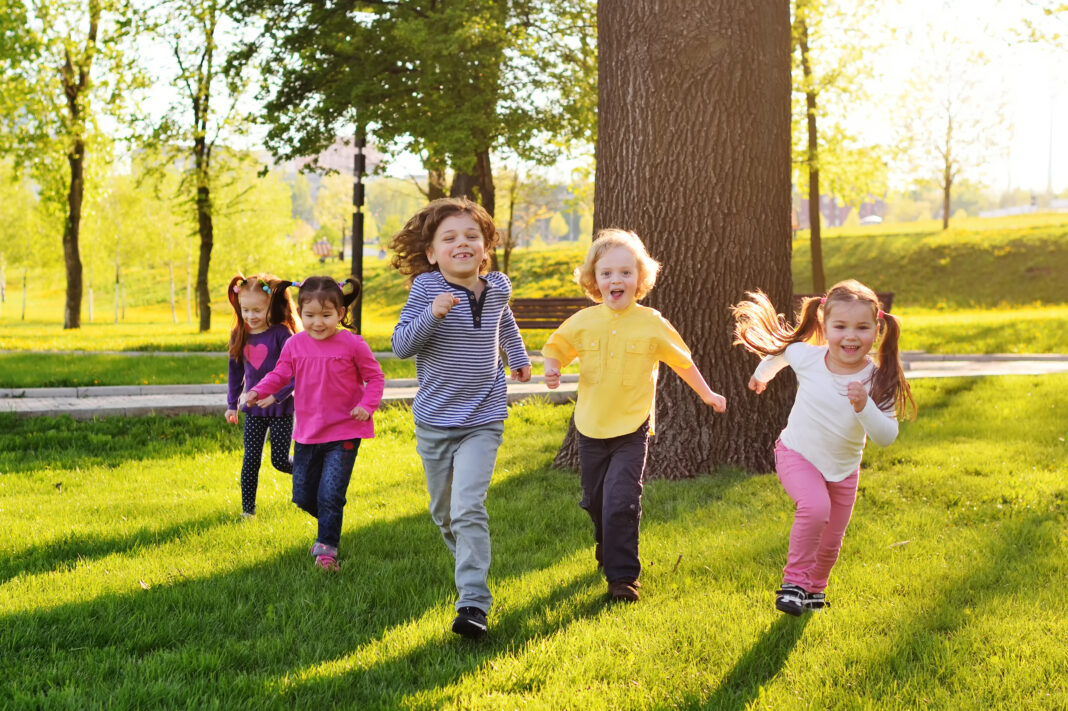 a group of small happy children run through the park in the background of grass and trees.