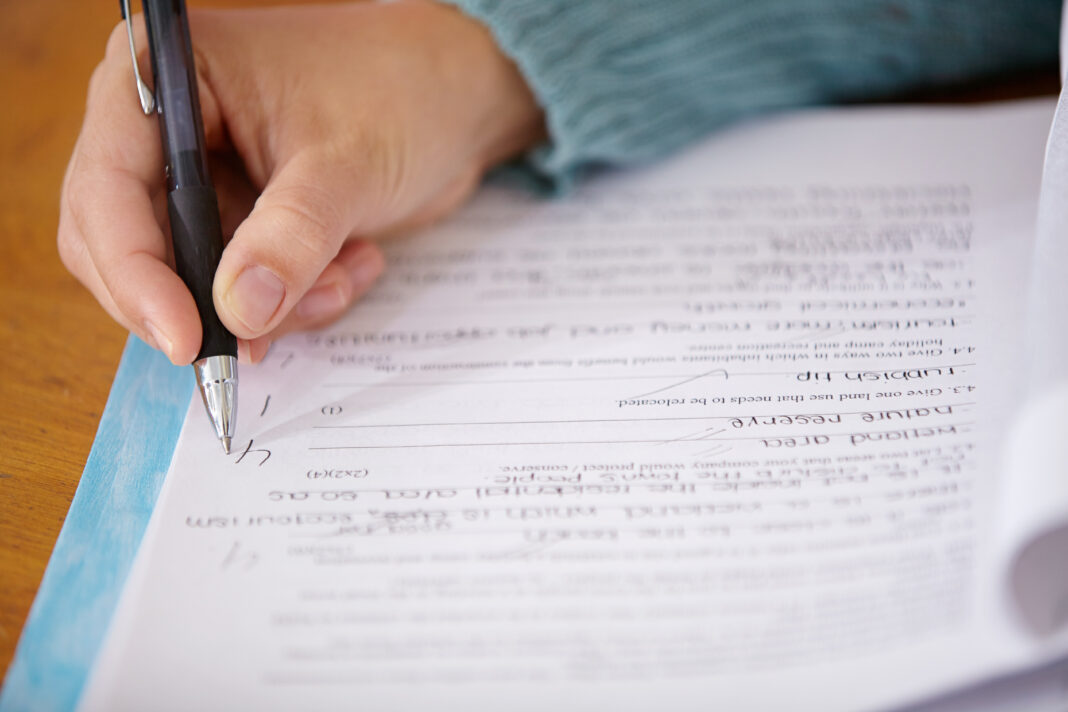 Hand, pen and teacher mark paper, education and learning with closeup of test or assignment on desk in classroom. Person in class, exam or project, grade and feedback with results, notes and writing