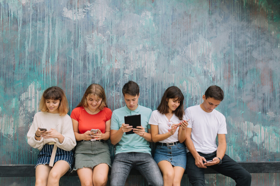 Group of teenagers sitting at the colored wall and using their mobile phones and gadgets