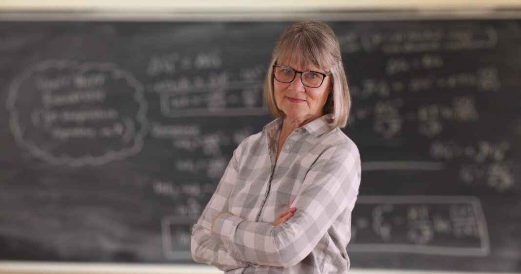 Confident senior woman teacher with arms crossed standing in front of chalkboard