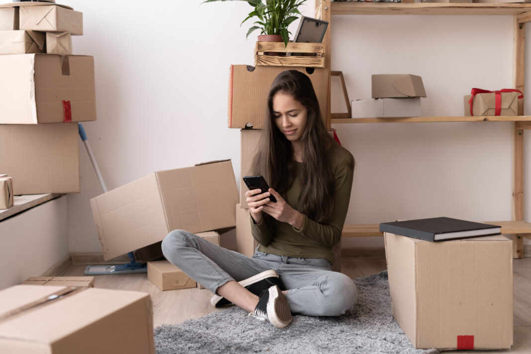 Happy young Hispanic woman using smartphone in living room at new house with stack of cardboard boxes on moving day